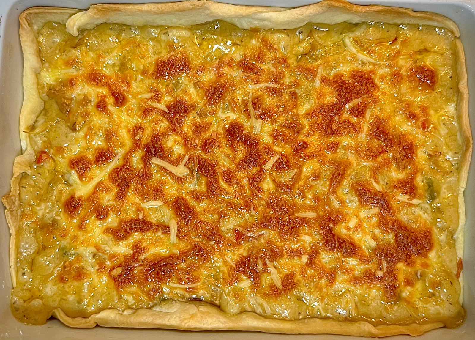 Foto desde arriba de un pastel de tofu recién sacado del horno. El pastel está en una fuente de horno blanca y cuadrada, se aprecian los bordes de hojaldre dorados y el centro con queso fundido en la superficie.